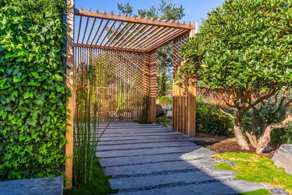 Pergola w ogrodzie z drewnianymi elementami przypominającymi deseczki do wnęk balkonowych.