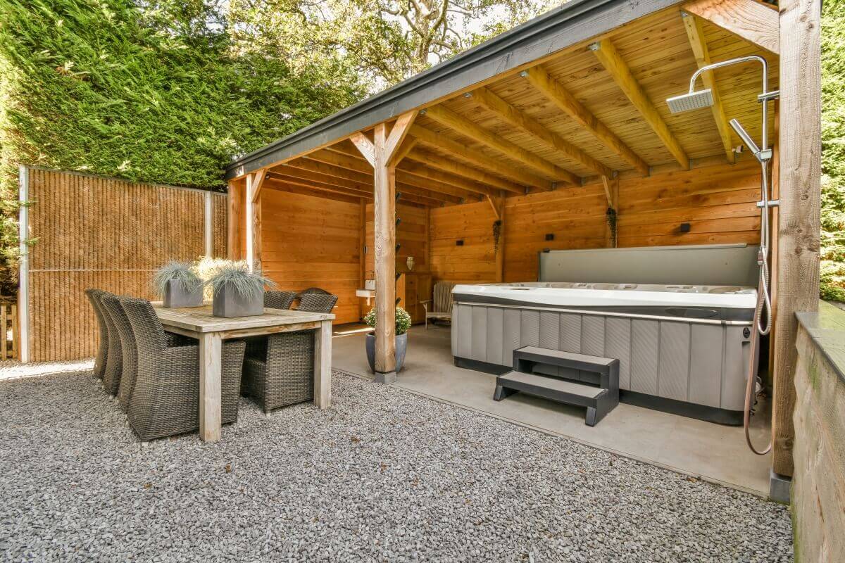 Covered gazebo with hot tub and table, surrounded by wooden plank buildings.